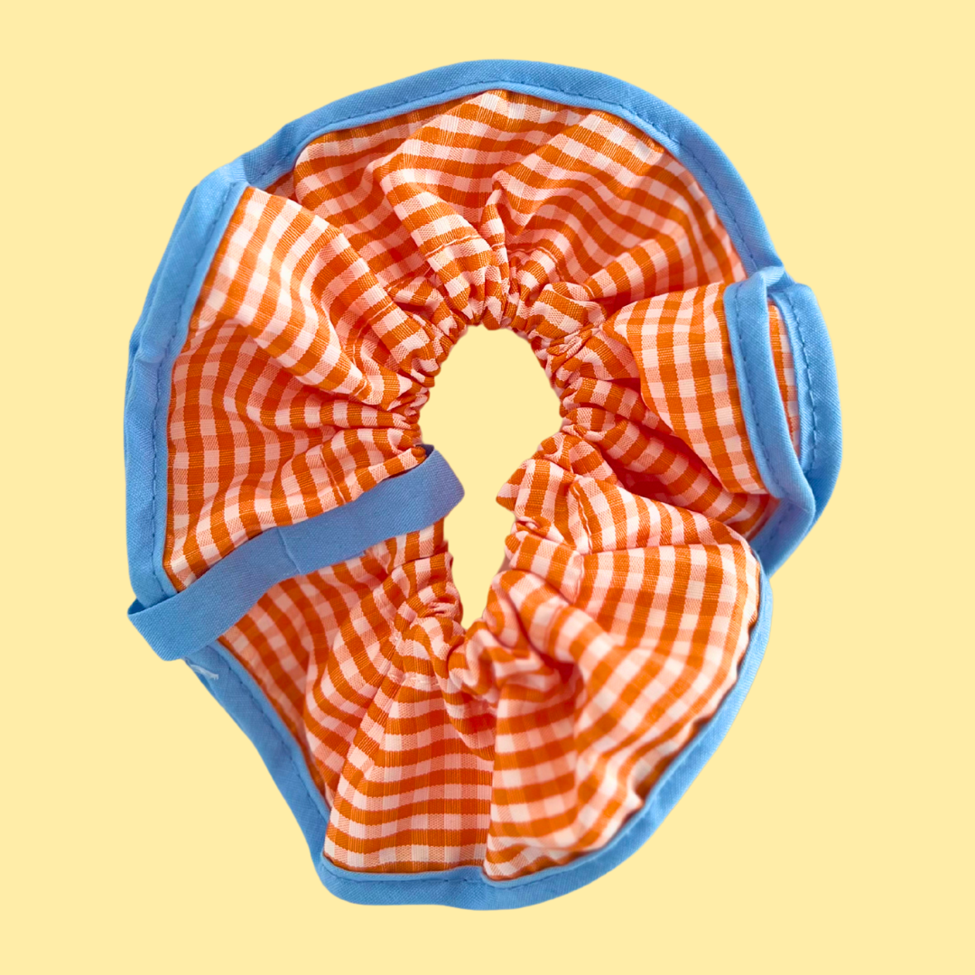 Scrunchie para pelo o muñeca con cuadros en tonos naranja y remates en azul de La Crème Organics. Accesorio ideal para outfits alegres y coloridos.