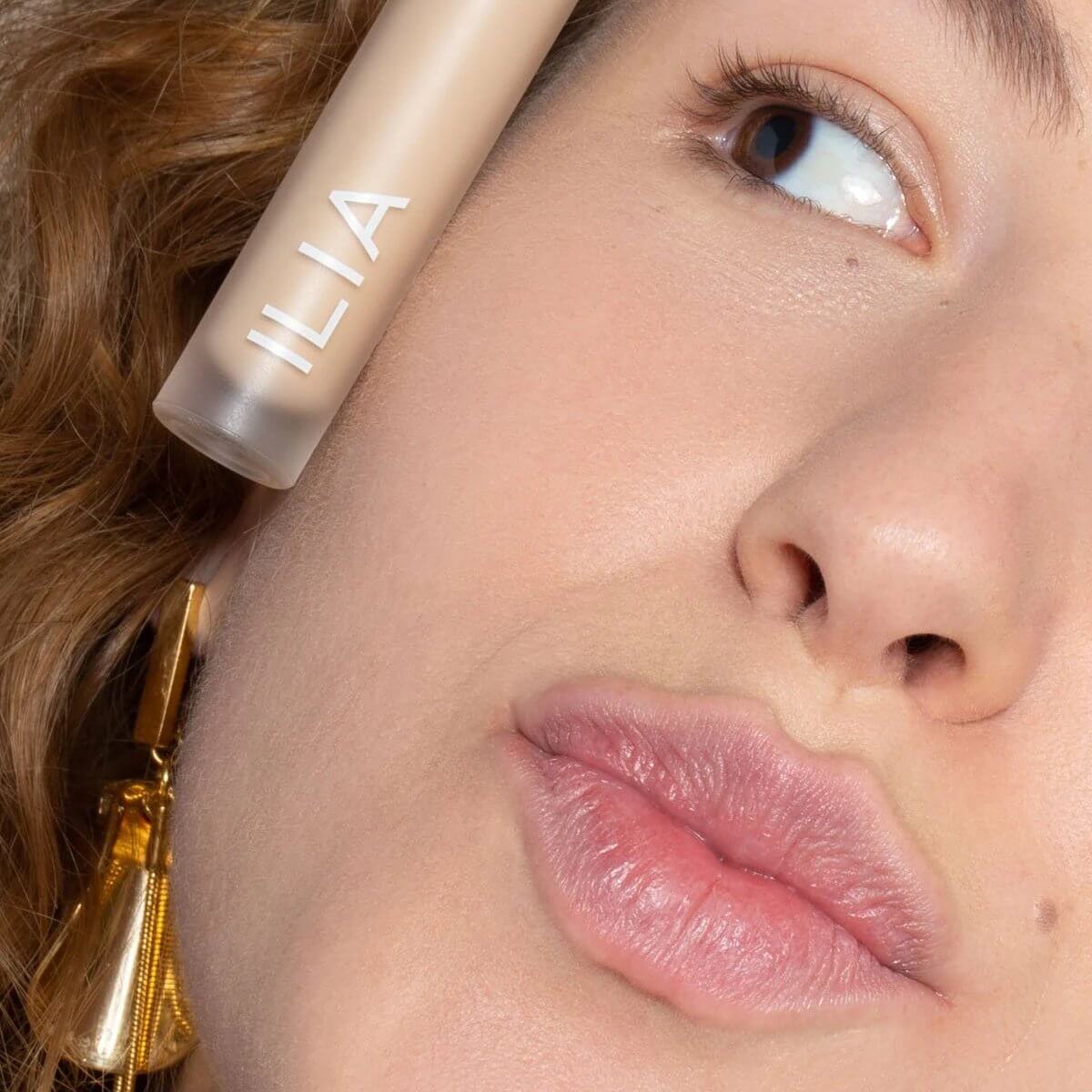 Rostro de mujer sonriendo con el corrector 'Chicory SC1' aplicado y el frasco de ILIA apoyado en la mejilla.